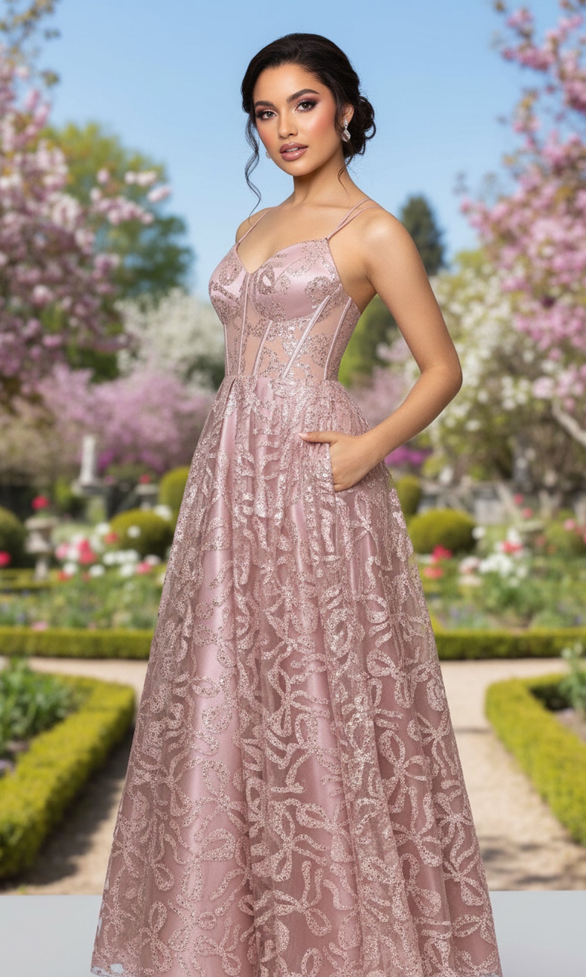 Wearing the Blondie Nites 6056BN long prom dress with a light pink glitter-print, fitted corset bodice, and flowing a-line skirt, a woman stands in a formal garden amid blooming trees and manicured pathways.